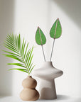 Two green stained glass mulberry leaf stems in a white vase beside a tan vase with a palm frond