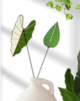 Green and pale stained glass leaf stems in a white jug against a white wall