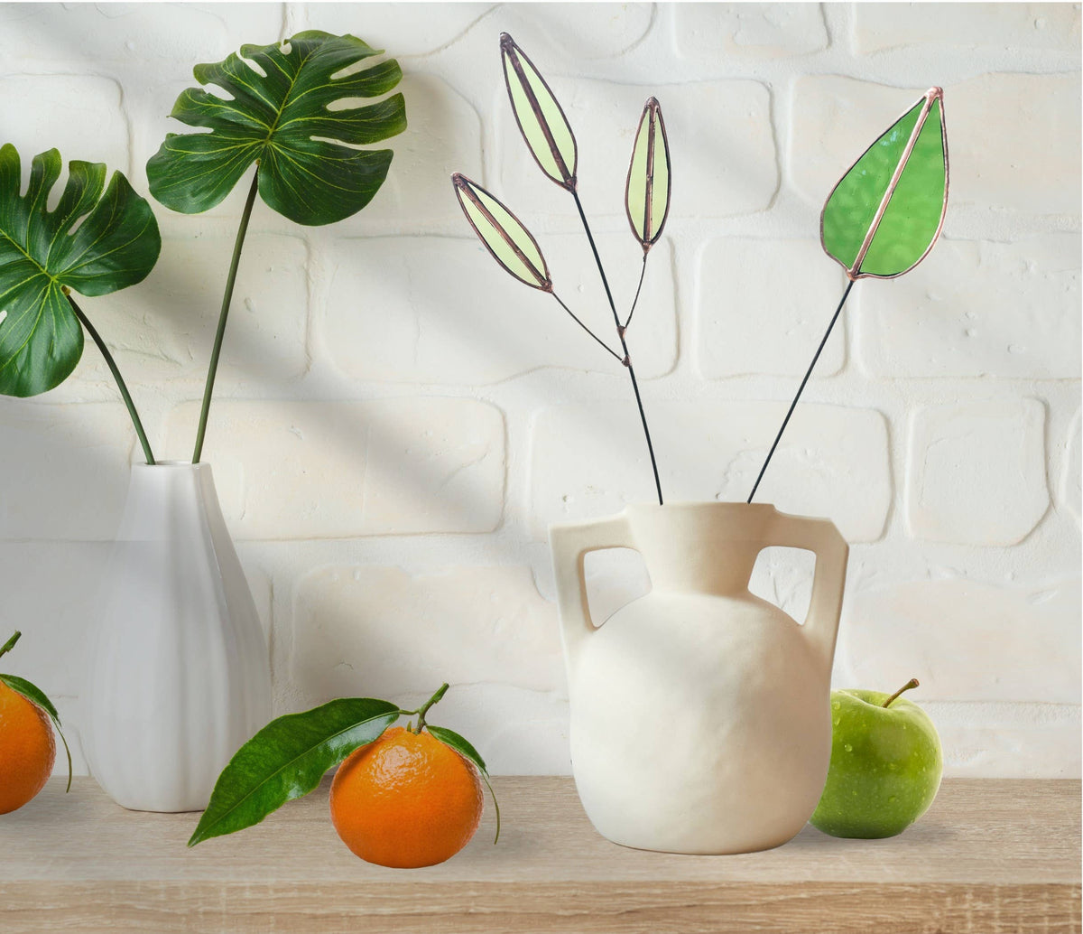Green mulberry leaf and pale seed pod stems in a white jug on a shelf with an orange and an apple