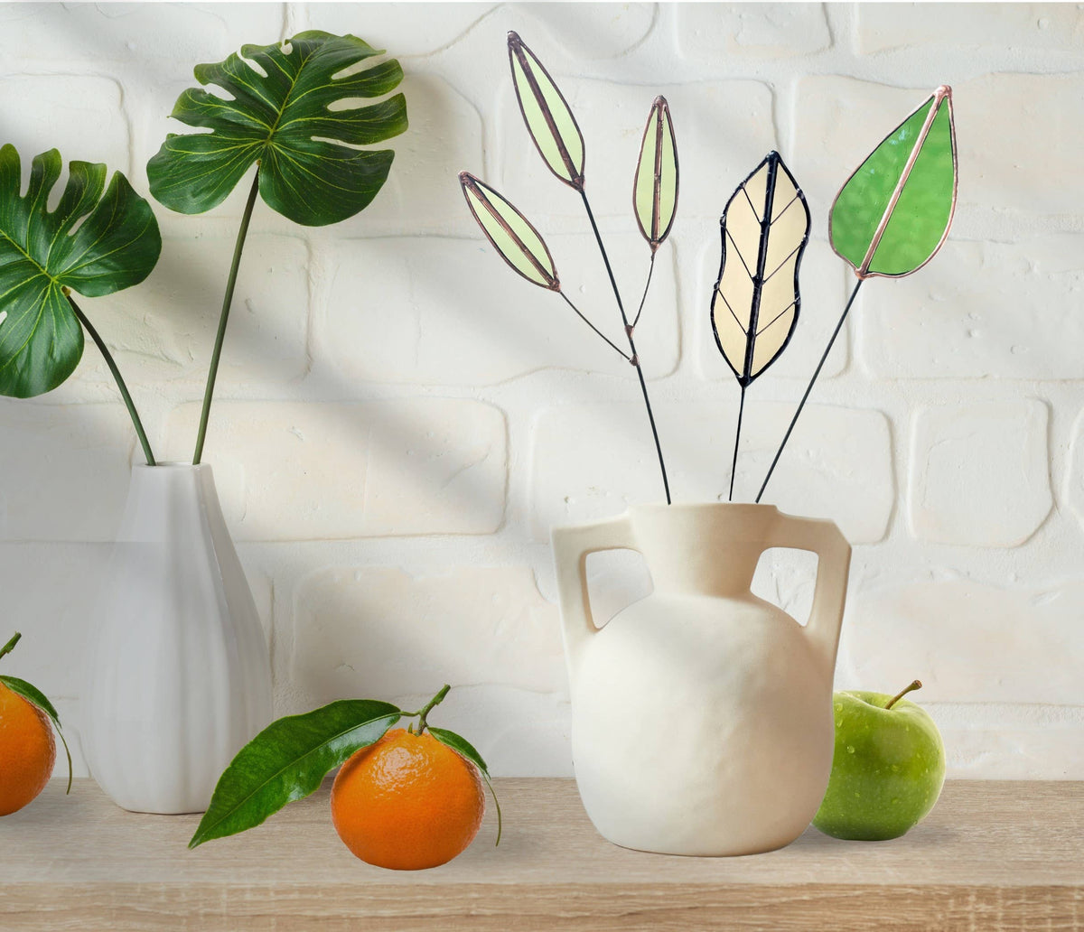 Green, pale and tan glass leaf stems in a white jug on a shelf with an orange and an apple