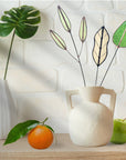Green, pale and tan glass leaf stems in a white jug on a shelf with an orange and an apple