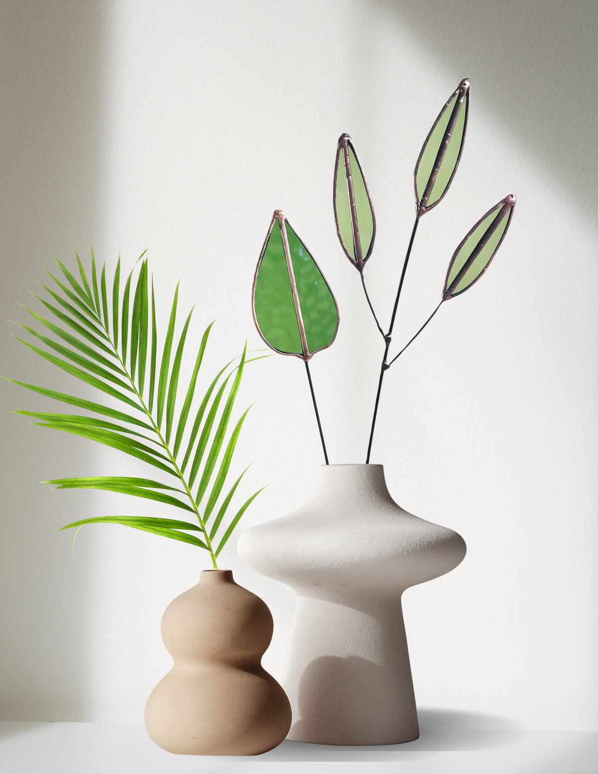 Green mulberry leaf stem with pale seed pod stems in a white vase beside a tan vase and palm frond