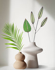 Green mulberry leaf stem with pale seed pod stems in a white vase beside a tan vase and palm frond