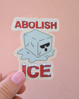 Sticker with 'Abolish Ice' text held by a hand against a pink background