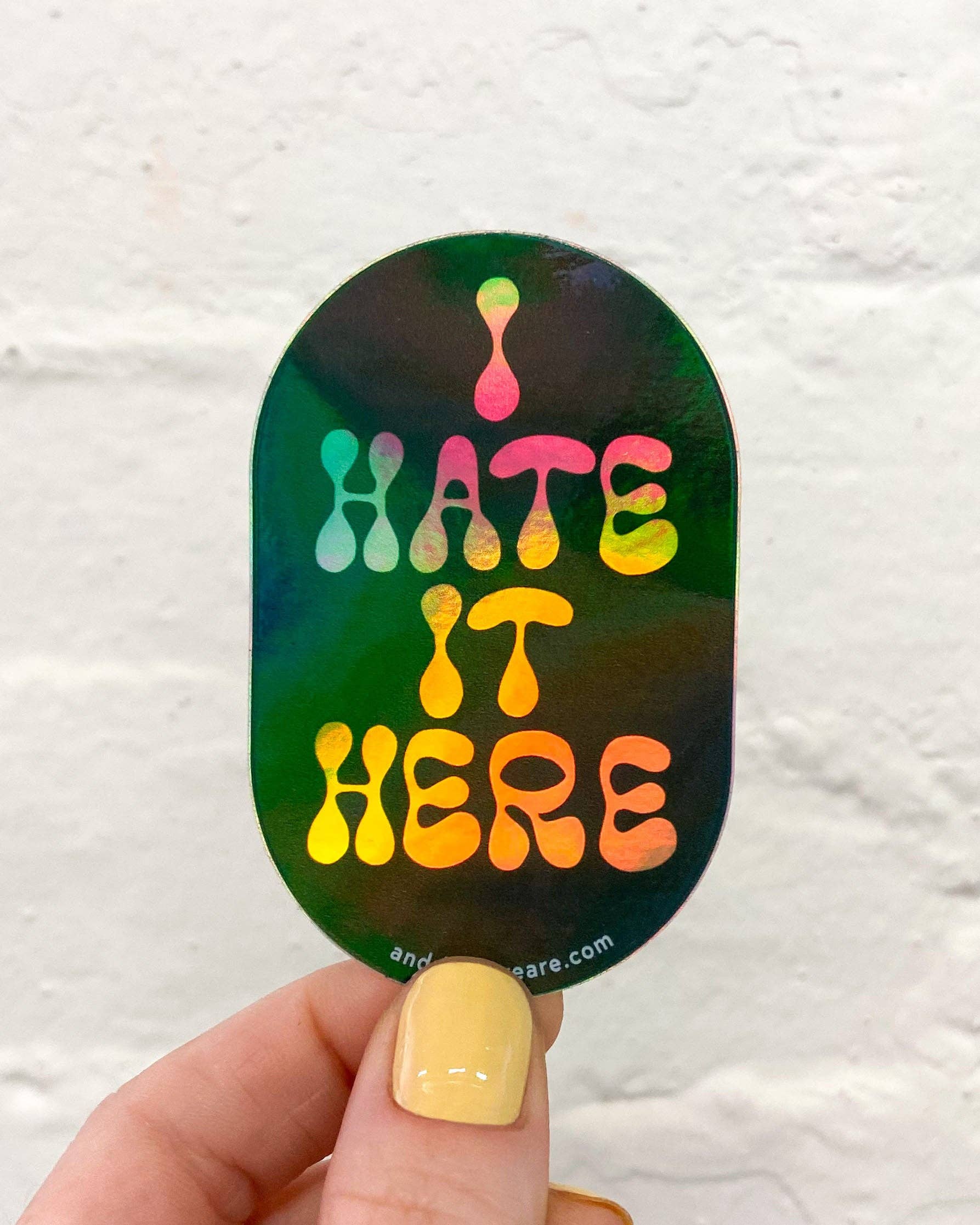 Sticker with 'HATE IT HERE' text held by a hand against a white background