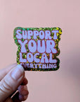 Holographic sticker with 'Support Your Local Everything' text held by a hand against a blurred background