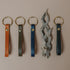 Four leather key fobs in tan, olive, indigo and black with brass rings beside driftwood