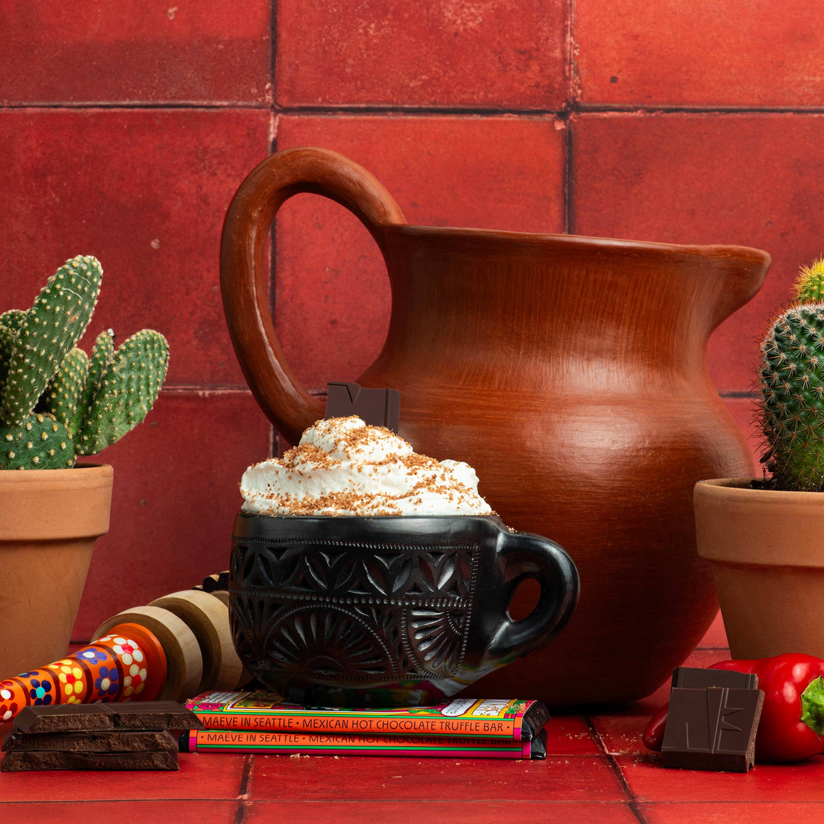Mug of hot chocolate topped with whipped cream next to a clay pitcher, cacti and Maeve Mexican Hot Chocolate bars on a red tile backdrop