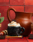Mug of hot chocolate topped with whipped cream next to a clay pitcher, cacti and Maeve Mexican Hot Chocolate bars on a red tile backdrop