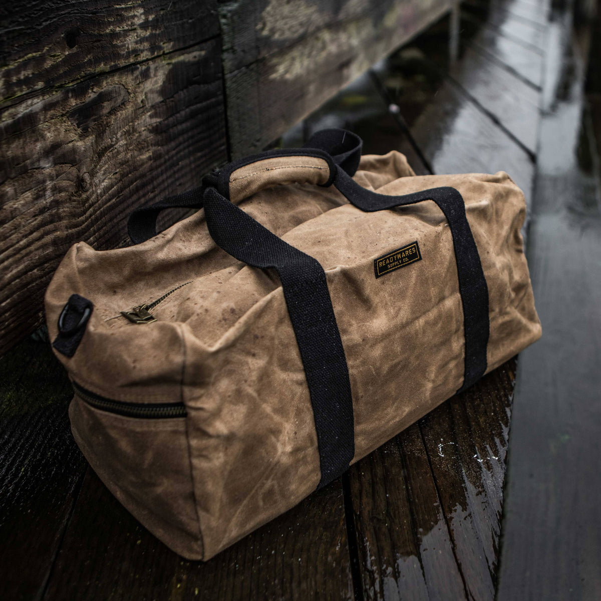 Tan waxed canvas duffel bag with navy straps on a wet wooden dock