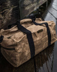 Tan waxed canvas duffel bag with navy straps on a wet wooden dock
