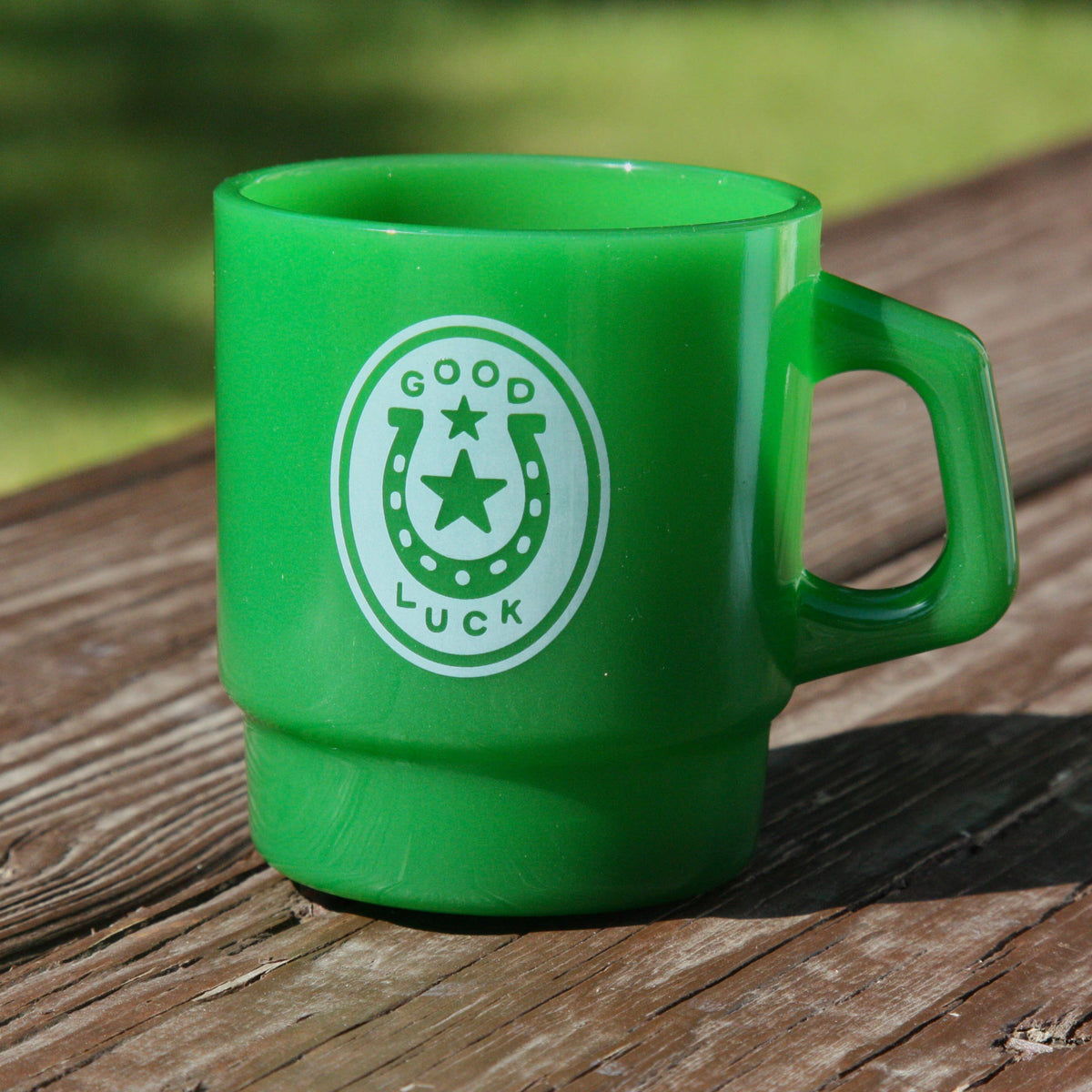 A bright green plastic mug with a white circular 'Good Luck' horseshoe graphic, sitting on a weathered wooden deck with a blurred green grass background.