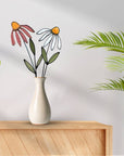 Pink and white stained glass coneflowers in a cream vase on a wood shelf with palm leaves