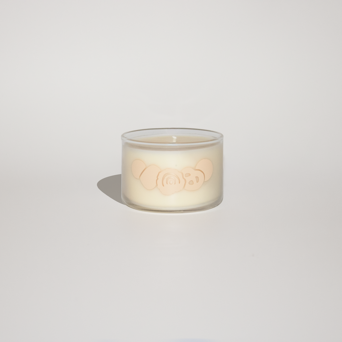 A small clear glass jar candle with creamy off-white wax and a pale peach embossed design on the front, photographed on a clean light gray background with a soft shadow.