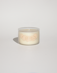 A small clear glass jar candle with creamy off-white wax and a pale peach embossed design on the front, photographed on a clean light gray background with a soft shadow.