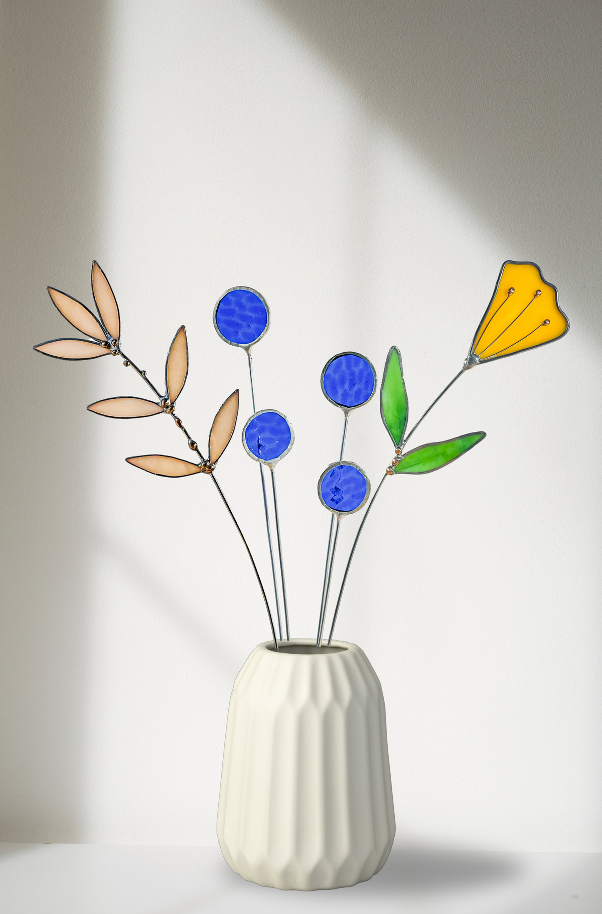 Blue thistles, pale leaf stems and an orange glass flower in a white ribbed vase in soft window light