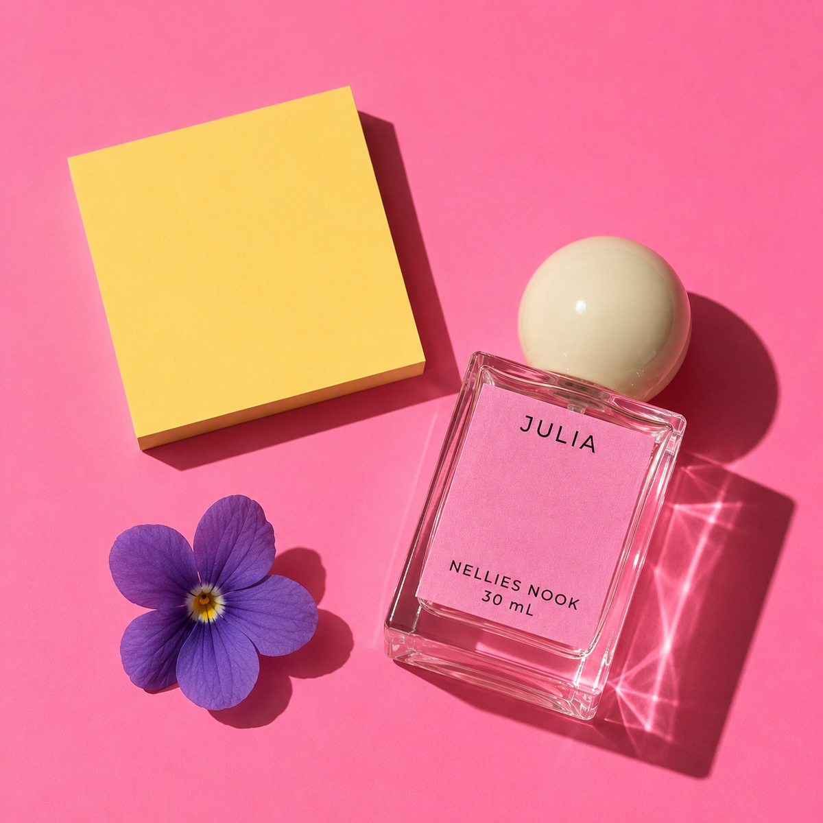 Julia perfume bottle beside a purple violet flower and a yellow block on a pink background