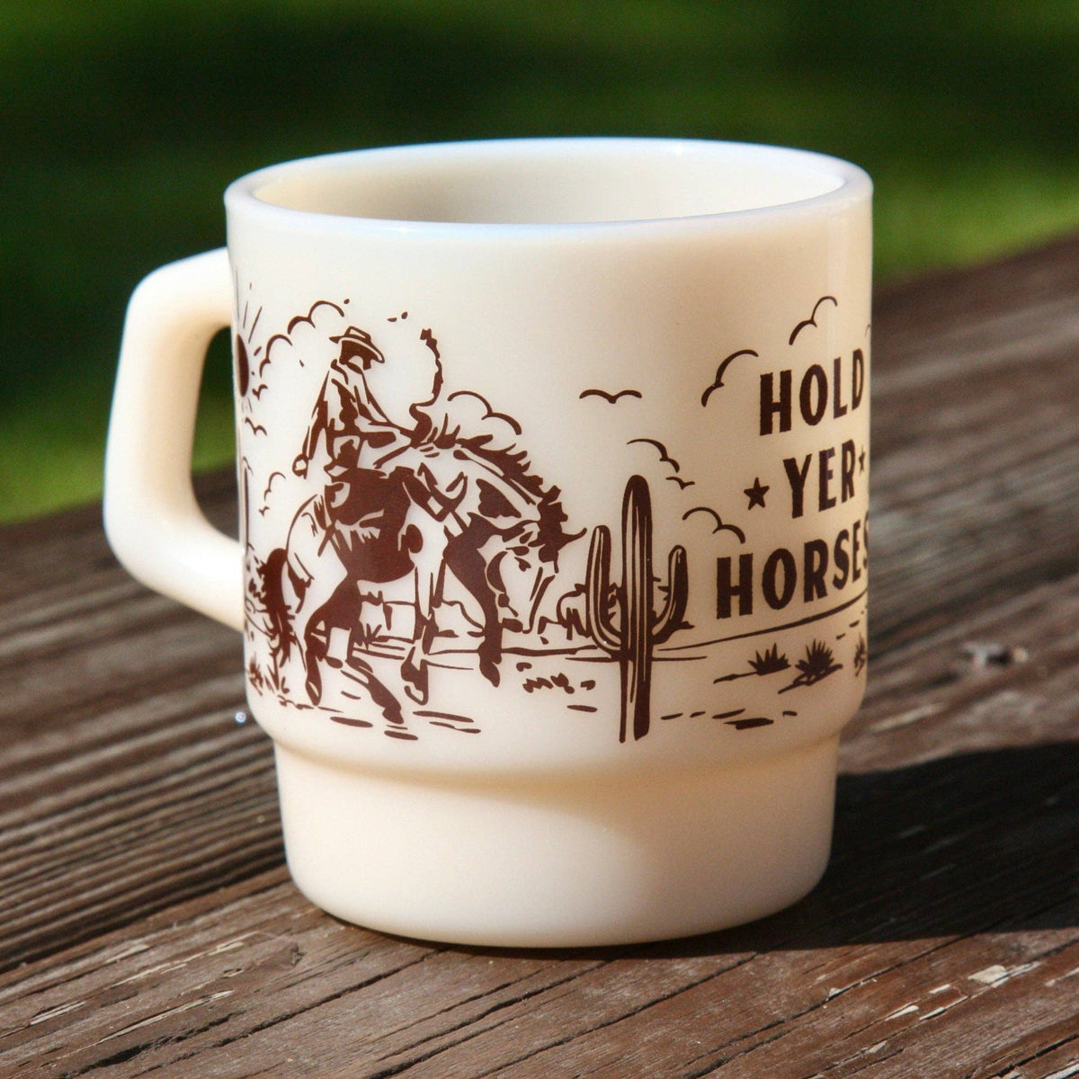 An off-white ceramic mug with a brown western-themed graphic and the text 'HOLD YER HORSES' sitting on a weathered wooden surface with a blurred green outdoor background.