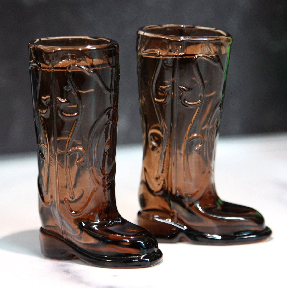Pair of brown glass cowboy boot shot glasses on a white surface with a dark background