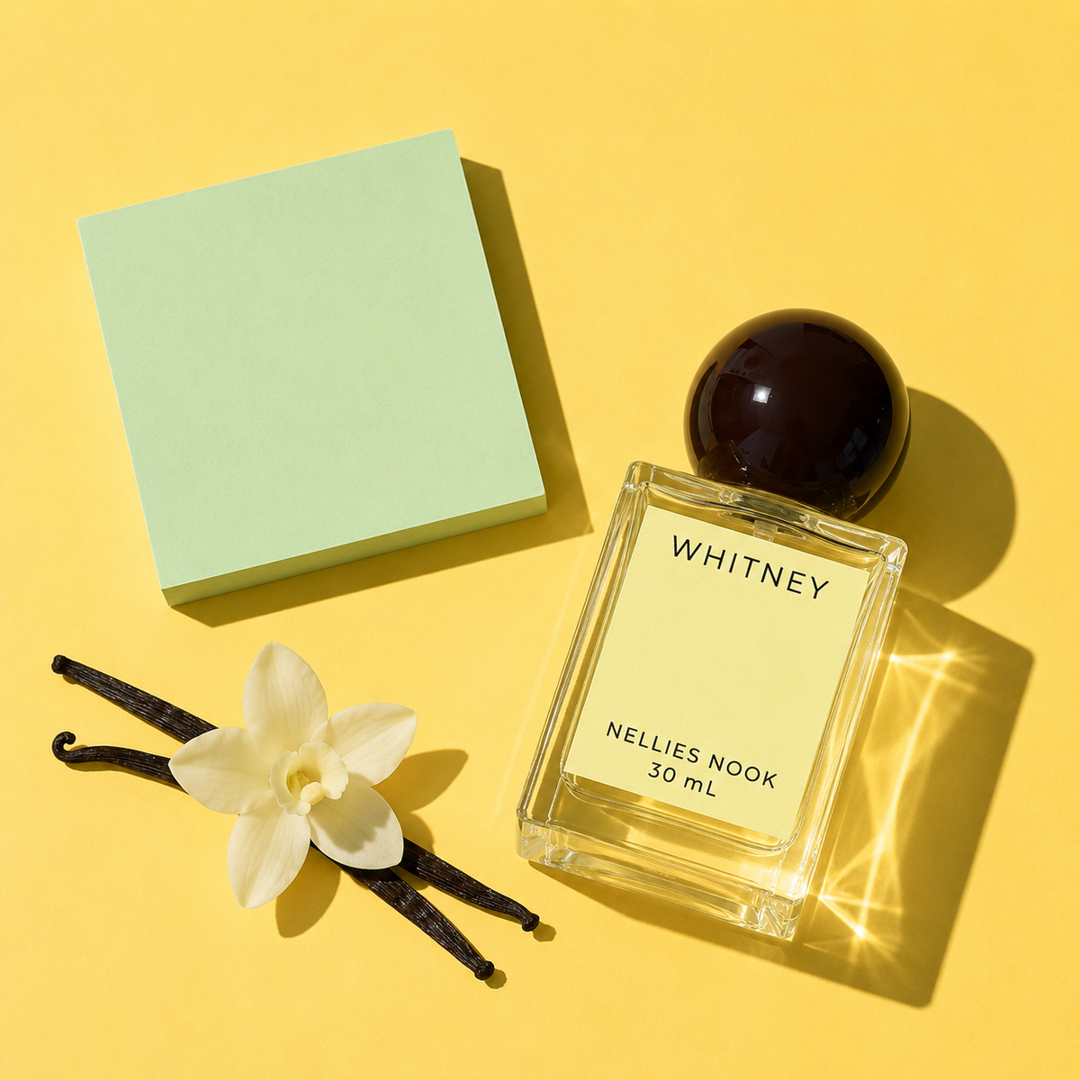 Whitney perfume bottle beside vanilla pods, a white flower and a mint green block on a yellow background