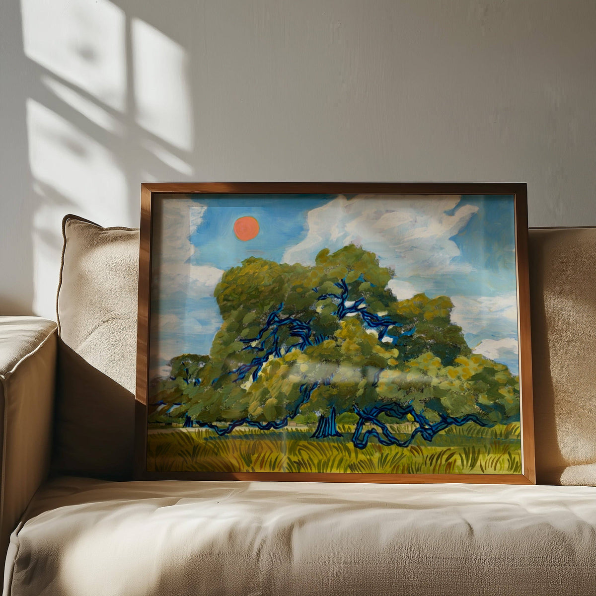 Framed live oak landscape print leaning against a beige sofa in a sunlit room