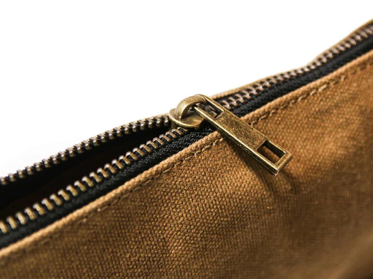Close-up of a brass zipper pull on tan waxed canvas, slightly unzipped