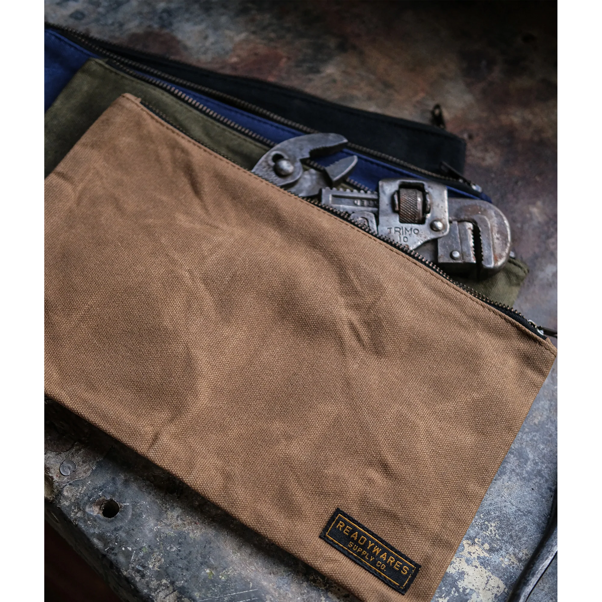 Tan waxed canvas tool bag over navy, olive and black bags with a pipe wrench on a weathered metal surface