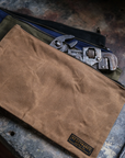 Tan waxed canvas tool bag over navy, olive and black bags with a pipe wrench on a weathered metal surface