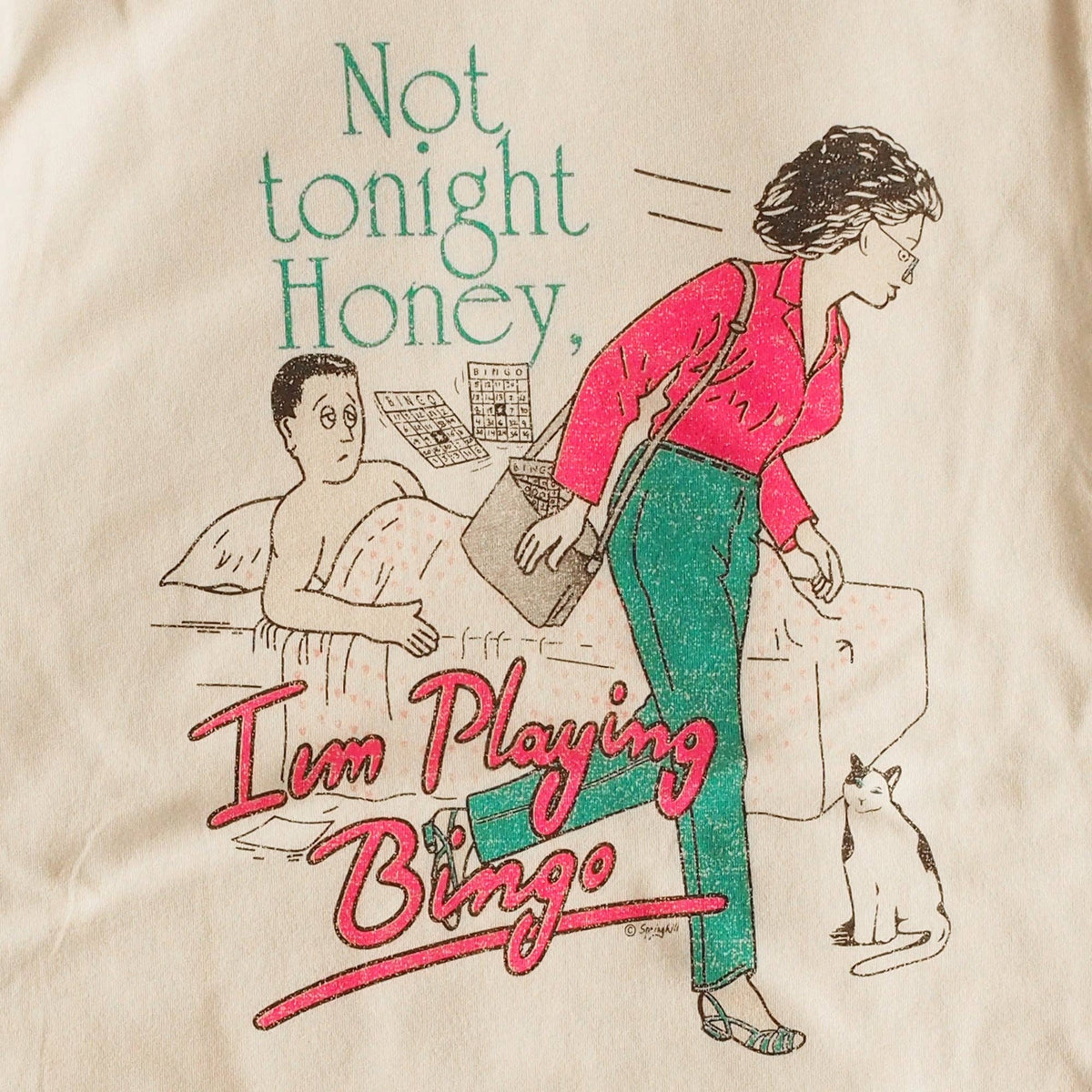 A close-up of a beige fabric surface printed with a retro-style illustration and the text 'Not tonight Honey, I'm Playing Bingo.' The print shows a woman in a pink top and green pants walking away from a bed, with a man in bed, a cat, and bingo cards.